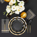 A black Sophistiplate wavy paper salad plate with gold trim and white flowers on a table set with black and gold paper dinnerware.