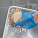 A person in blue gloves cleaning a metal tray on a counter with a Carlisle Sparta tan round palm scrub brush.
