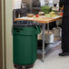A person standing next to a green Rubbermaid commercial trash can.