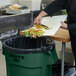 A person throwing food into a Rubbermaid green round trash can.