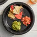 A black Ecopax polypropylene plate with chips and guacamole on a table with a plate of chips and salsa.
