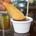 A person holding a fried chicken stick over a small white Thunder Group ramekin of sauce.