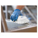 A hand in a blue glove using a Vikan white scrub brush to clean a metal surface.