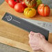 A hand holding a black Mercer Culinary polypropylene knife blade guard over a tomato on a cutting board.