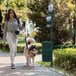 A woman walking a dog next to a green Flash Furniture pet waste station with a round lidded trash can.