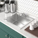 A stainless steel Regency drop-in sink with a faucet and white mugs and containers.
