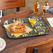 A rectangular Cambro tray with a beer design holding a pretzel and a yellow container with a white rim on it.