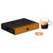 A black and orange Nespresso Professional Caffe Caramello coffee box on a table with a glass of brown coffee.