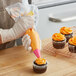 A person wearing gloves holds a Wilton plastic pastry bag filled with orange frosting over a cupcake.