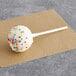 A Coco Bakery birthday cake pop with white frosting and colorful sprinkles on a stick.