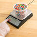 A hand using a Taylor digital portion scale to weigh a container of sprinkles.