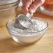 A close-up of a hand using a metal cup to scoop De Tulpen Tapioca Starch into a glass bowl.