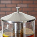 A Vollrath stainless steel lid with a chrome handle on a glass beverage dispenser filled with brown liquid and lemon slices.