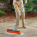 A person using a Seymour Jobsite rough surface street push broom with a blue handle to sweep a sidewalk.
