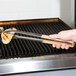 A person using Vollrath Jacob's Pride stainless steel tongs with tan handles to cook on a grill.
