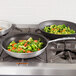 Two Vollrath Wear-Ever non-stick fry pans with vegetables and meat cooking on a stove.
