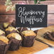 A group of blueberry muffins in brown wrappers on a wood surface with mini chalk cards on a table