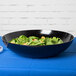 A black GET Siciliano bowl filled with green leaves on a blue table.