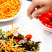 A hand using a Fineline clear plastic tong to serve salad with carrots and tomatoes.
