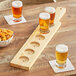 An Acopa natural wood flight paddle with a row of beer glasses and a bowl of crackers on a table.