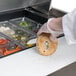 A hand in a glove is using a knife to cut a bagel on a stainless steel counter.