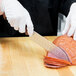 A person wearing white gloves cutting ham with a Mercer Culinary Millennia Colors Santoku knife.