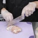 A person in gloves uses a Mercer Culinary purple chef knife to cut raw chicken on a purple surface.