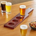 An Acopa mahogany beer flight paddle on a table with glasses of beer and a bowl of mixed nuts.