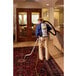 A man using a ProTeam QuietPro backpack vacuum to clean a carpet.
