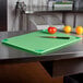 A San Jamar green cutting board on a counter with a knife and a tomato.