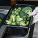 A gloved hand holding a black Vollrath Super Pan container of broccoli.