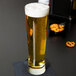 A Libbey Pinnacle pilsner glass of beer on a black surface with pretzels.