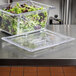 A Vollrath clear plastic lid on a container of green and red lettuce on a counter.