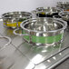 A yellow and silver Bon Chef steam table pot with riveted handles on a metal surface with a group of other Bon Chef pots.