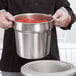 A person holding a Vollrath stainless steel pot of red sauce.