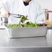 A chef using tongs to serve salad from a Vollrath stainless steel deli pan.