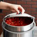 A hand with a Vollrath Kool Touch stainless steel hinged cover opening a pot of tomatoes.