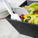 A salad in a black American Metalcraft square melamine bowl with a pair of tongs.