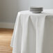 A white Intedge round poly cotton tablecloth on a table with a stack of white plates.