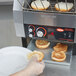 A person's hand in a plastic glove putting a piece of white bread in a Hatco TQ-800H conveyor toaster.