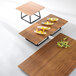 An Elite Global Solutions faux bamboo melamine serving board on a table with a bowl of fruit and wooden trays.