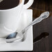 A Oneida Rosewood stainless steel demitasse spoon on a saucer next to a cup of coffee.