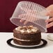 A hand reaching for a chocolate cake in a D&W Fine Pack plastic cake container with a clear dome lid.