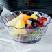 A clear plastic swirl bowl filled with fruit with a spoon next to it.