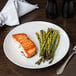 A white Siciliano plate with a piece of cooked salmon and asparagus on a table.