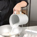 A person pouring milk from a white Vollrath aluminum measuring cup into a bowl.
