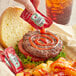 A hand holding a Heinz ketchup packet next to a burger.