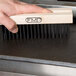 A hand uses an FMP grill cleaning brush head on a wooden stick to clean a grill.