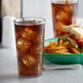 A Carlisle clear plastic tumbler filled with brown soda and ice on a table with a plate of french fries and a sandwich.