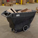 A large black Rubbermaid tilt truck trash cart full of boxes.